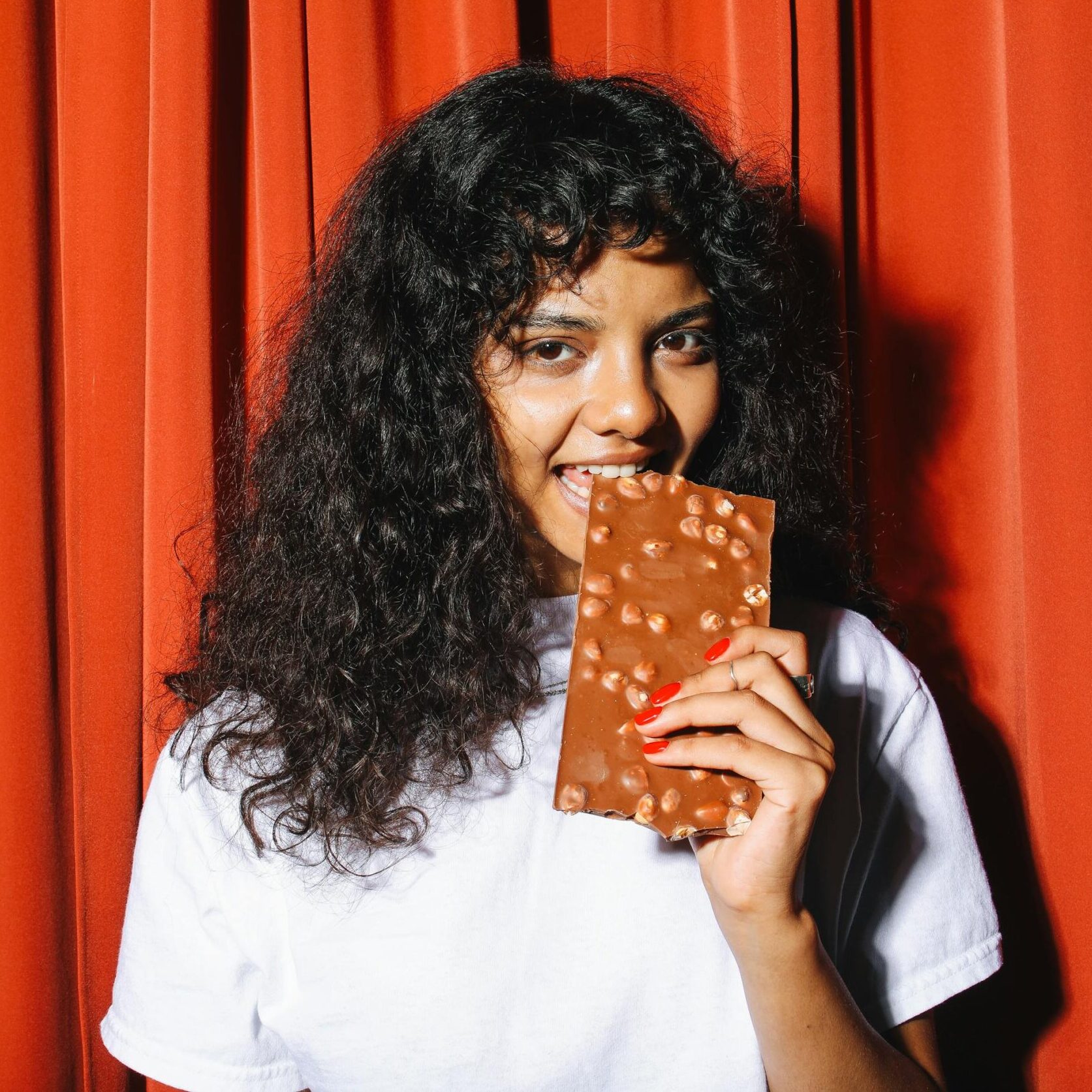 A woman with curly hair eats a chocolate bar in front of a red curtain, smiling.